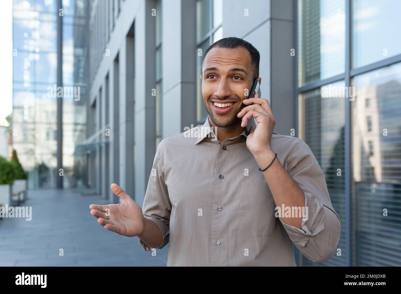 Allegro e sorridente lavoratore di ufficio afroamericano che parla al telefono, uomo fuori ufficio edificio in camicia a piedi in città, uomo d'affari in pausa pranzo. Foto Stock