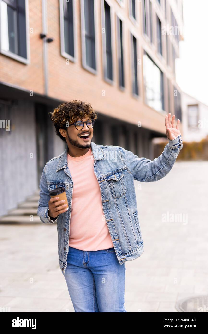 Vita in su ritratto dell'uomo indiano è in piedi con caffè per andare e salutare qualcuno con la mano Foto Stock