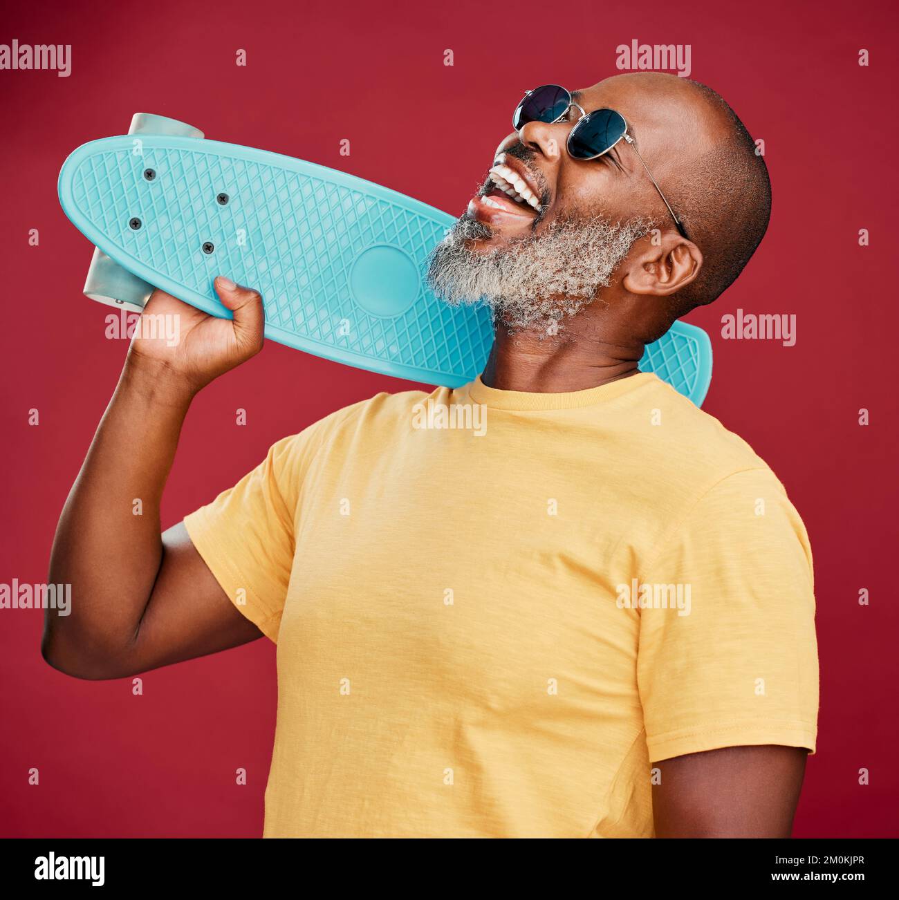 Un uomo afroamericano maturo in piedi con uno skateboard mini in studio isolato su uno sfondo rosso. Uomo bello e spensierato Foto Stock