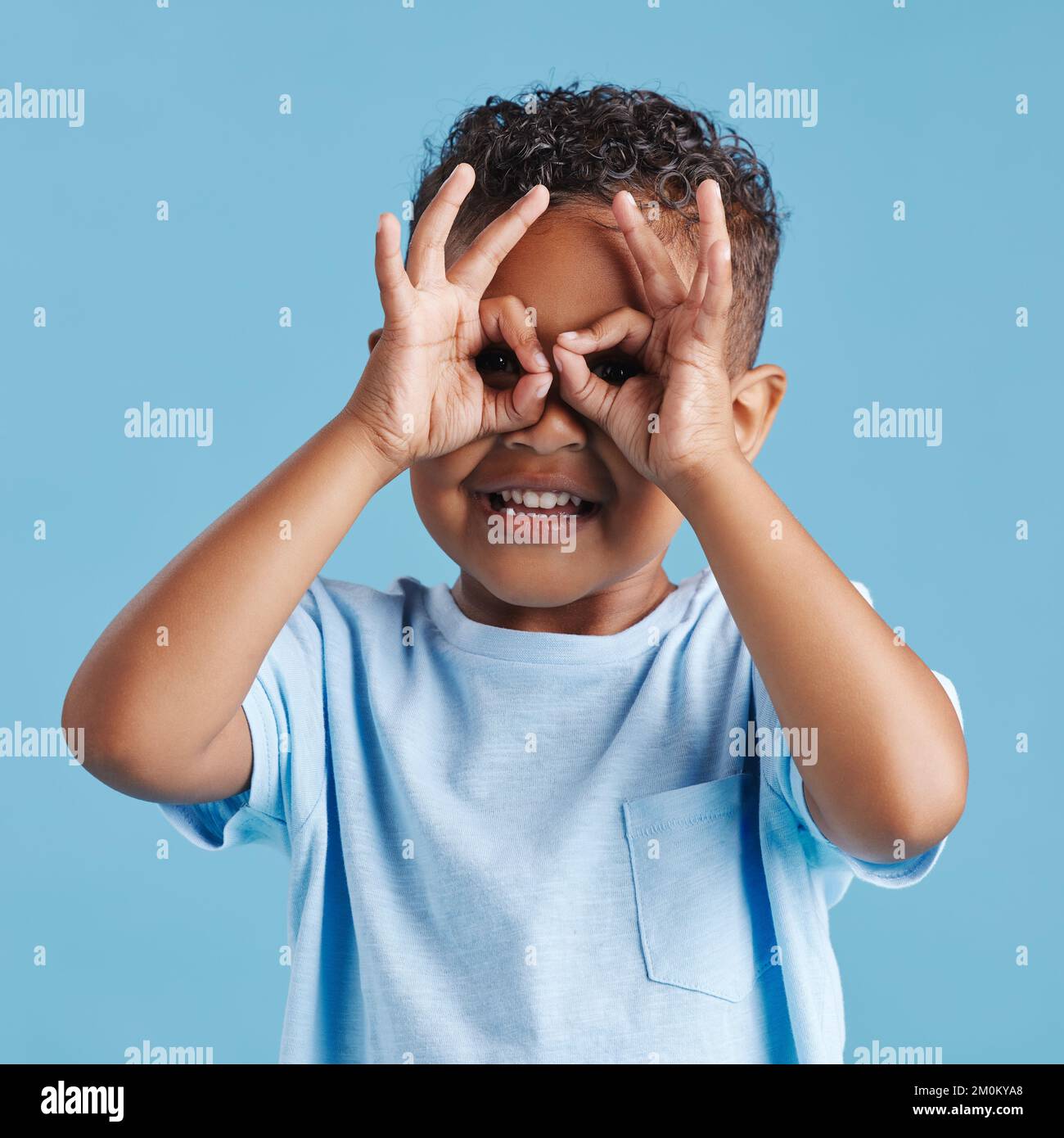 Ritratto di un ragazzino curioso che guarda attraverso le dita a forma di binocolo contro uno sfondo blu da studio. Bambino curioso esplorare Foto Stock