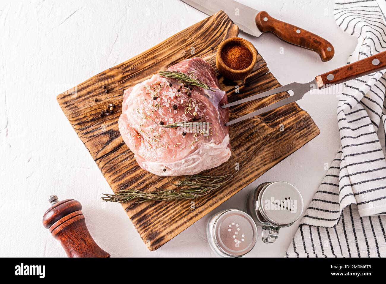 Un grosso pezzo di prosciutto di maiale si trova su un tagliere di carne di legno testurizzato con ingredienti per la cottura. coltello, forchetta per carne, spezie Foto Stock