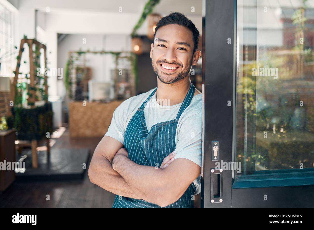 Ritratto di un giovane cameriere ispanico felice in piedi con le braccia incrociate alla porta di un negozio o caffè. Uomo cordiale e proprietario di una caffetteria Foto Stock