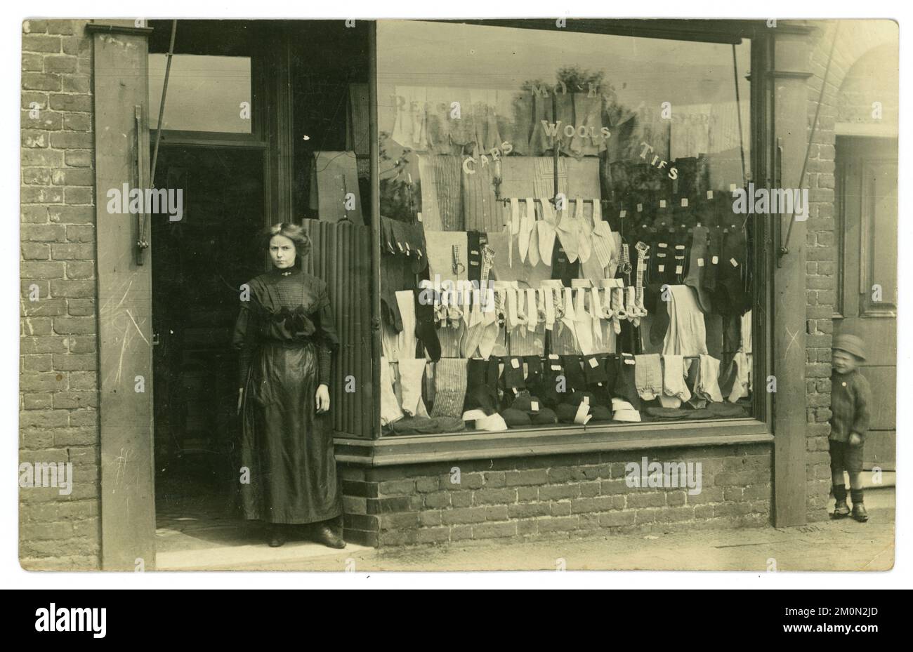 Originale affascinante post edoardiano / Titanic era cartolina del proprietario signora o assistente di vendita in piedi fuori di un negozio di abbigliamento maschile, nella finestra di cui pubblicizza le vendite di cravatte, camicie, lane, calze. C'è un bambino di un vicino che sbirciava da una porta dietro l'angolo. Un classico "moment in time" catturato. La cartolina è datata / pubblicata il 31 maggio 1911 da Warrington, Cheshire, Inghilterra, Regno Unito Foto Stock