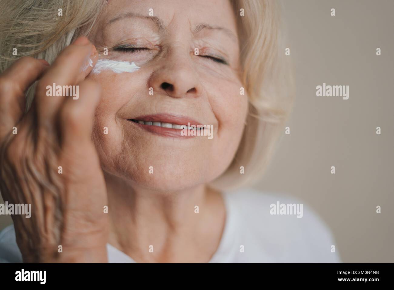 Donna matura felice che applica lozione anti-invecchiamento per rimuovere i cerchi scuri sotto gli occhi. Prodotto per il trucco. Sorridi il viso. Foto Stock