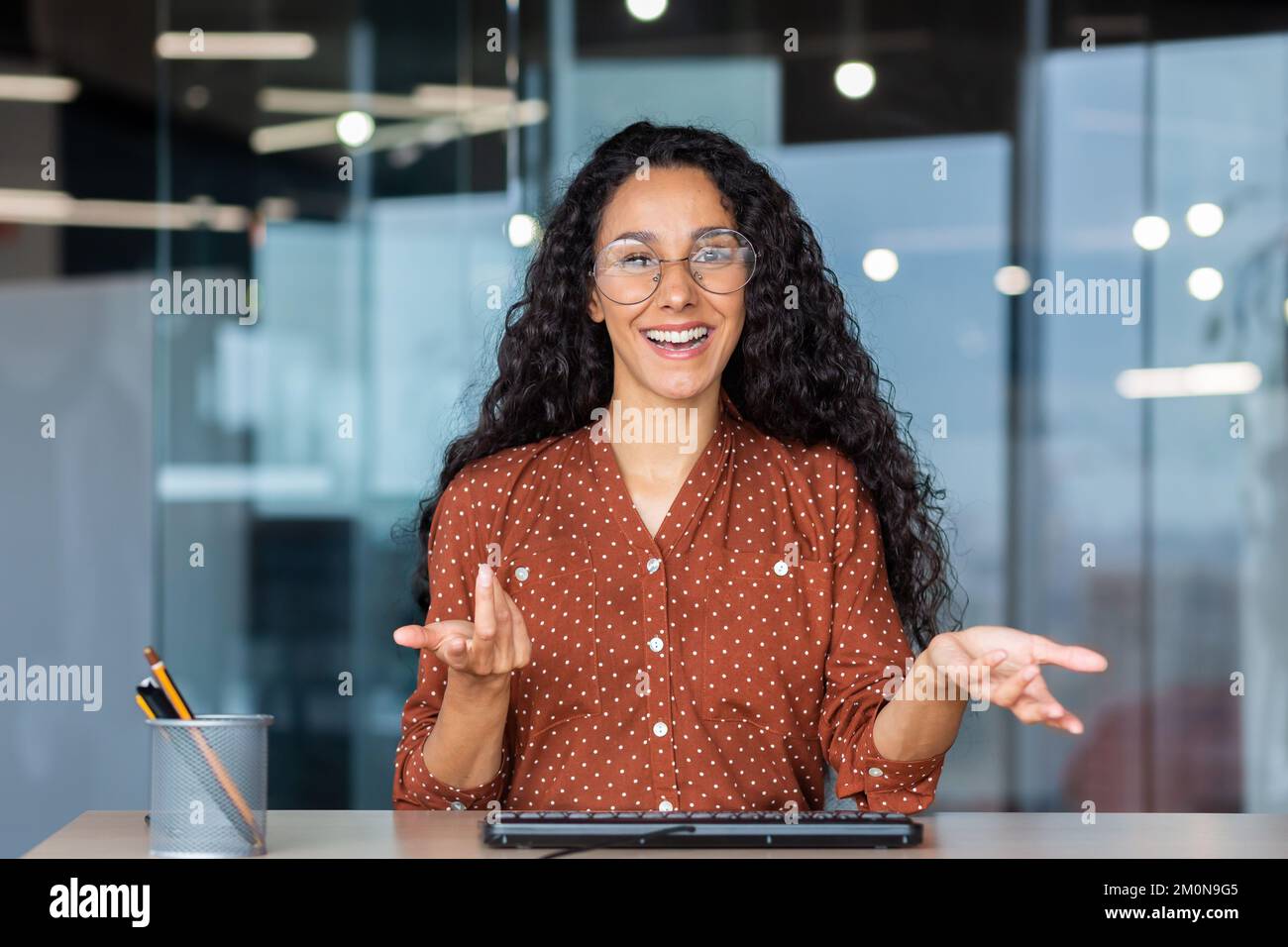 Assistenza clienti online videochiamata, donna latino-americana che guarda la webcam sorridendo e consultando i clienti, assistente che lavora all'interno dell'ufficio. Foto Stock