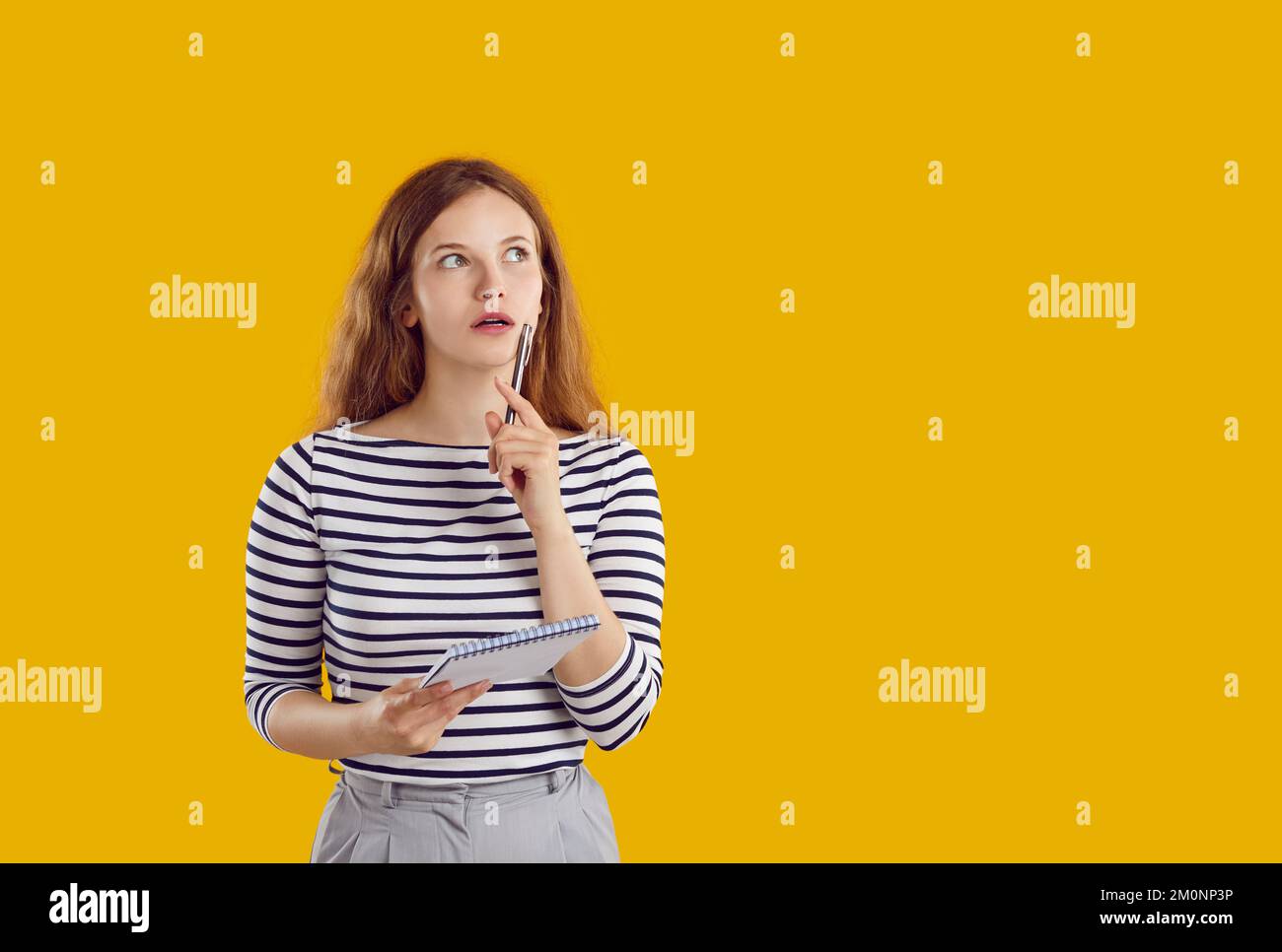 Donna in camicia a righe sta pensando di tenere la penna e il notebook guardando su sfondo giallo. Foto Stock