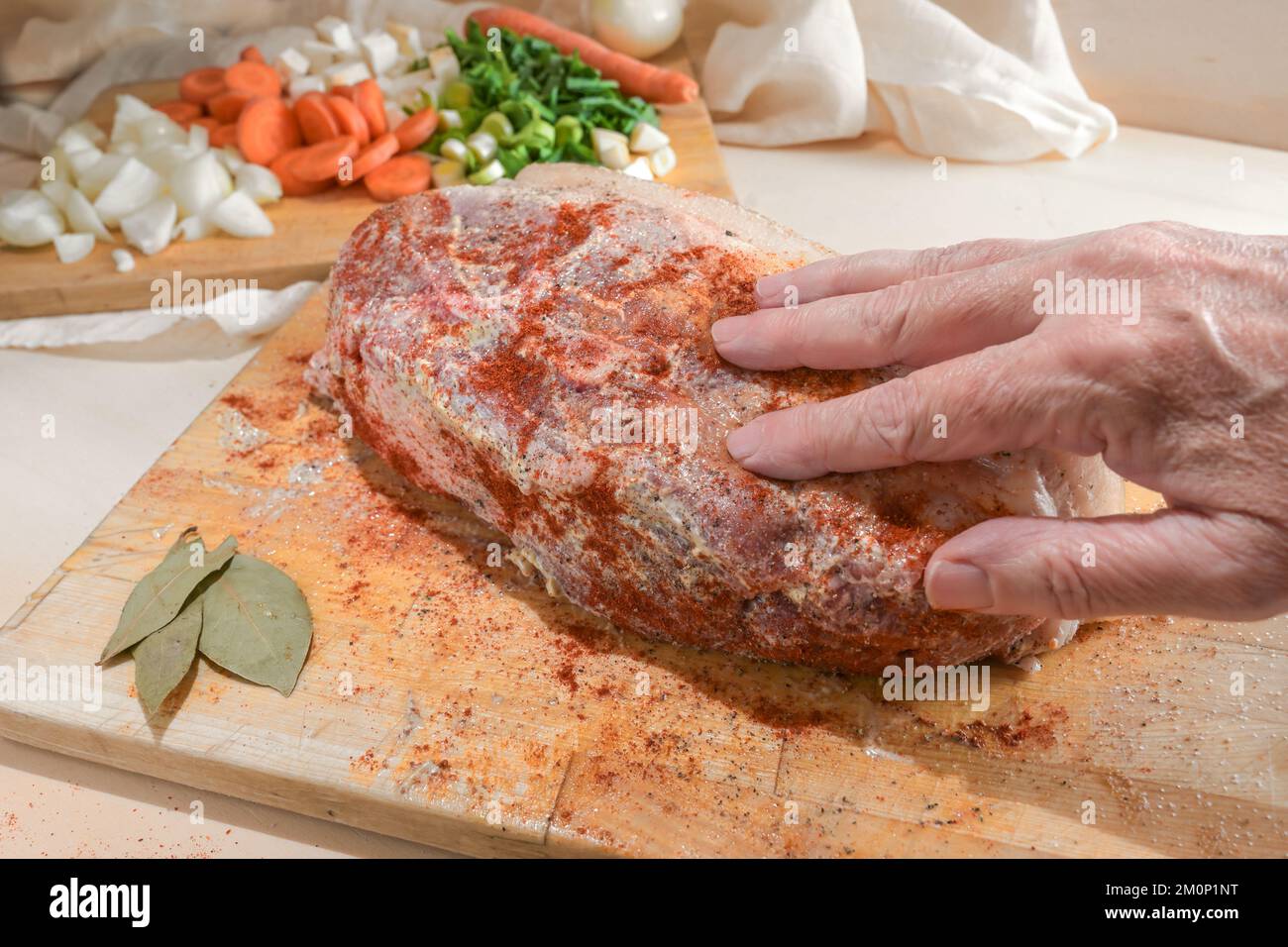 La mano strofina un pezzo di carne di maiale cruda con senape e spezie su un tagliere di legno, preparazione per un delizioso arrosto, fette di verdure nel b Foto Stock