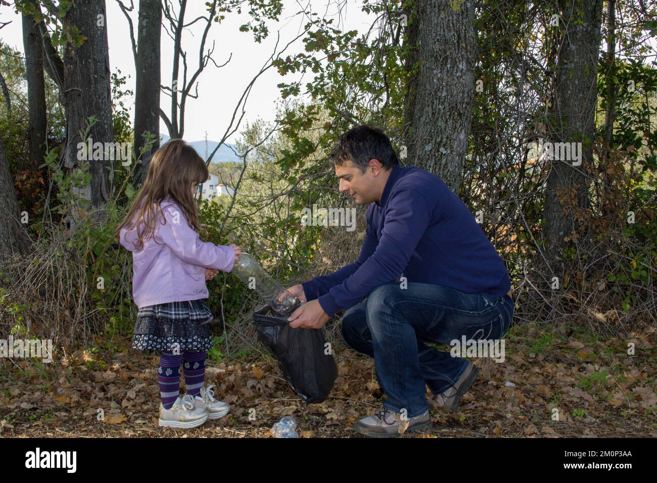 Immagine di un padre e di una figlia sorridenti mentre puliscono un boschetto di rifiuti e bottiglie di plastica. Famiglia rispettosa dell'ambiente e del riciclaggio Foto Stock