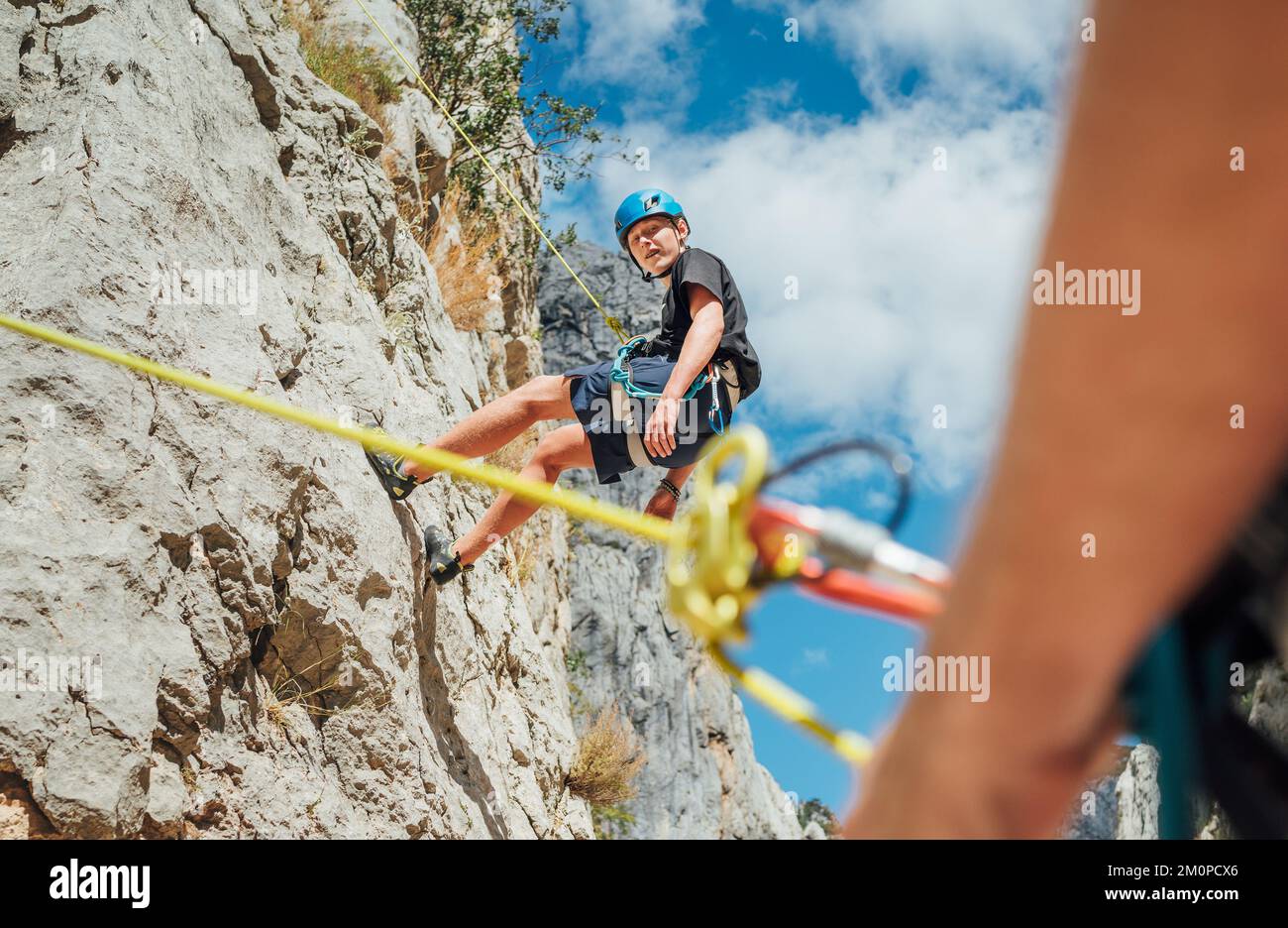 Ragazzo adolescente sulla parete di arrampicata naturale scogliera. Ragazzo che si aggrappa su una corda con un'imbracatura da arrampicata e il padre che lo cinge a terra con una bela Foto Stock