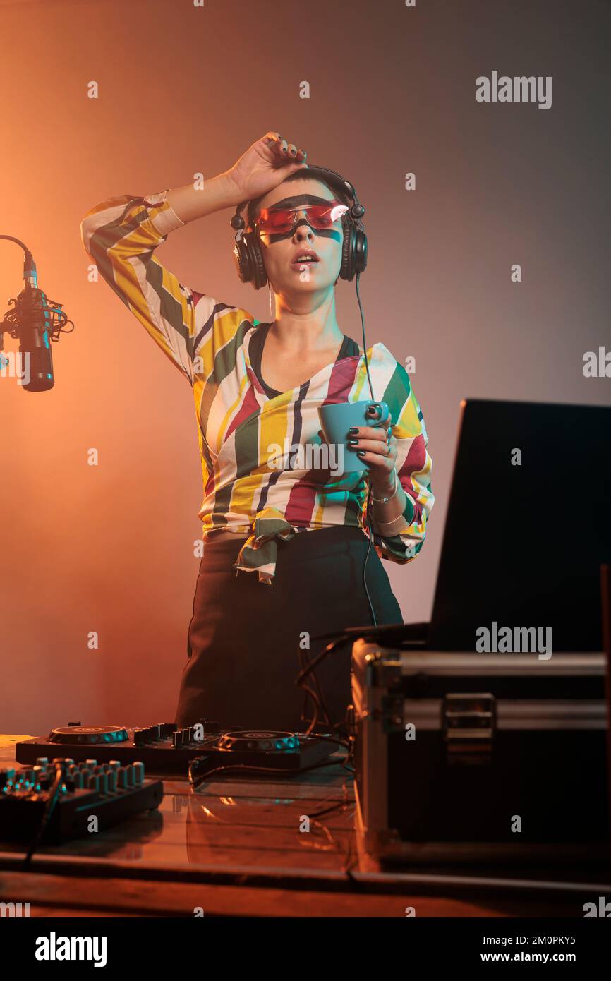 Stanco artista femminile bere caffè e sbadiglio, sensazione di esaurimento dopo la festa nightclub mescolando musica techno. Sonnolenza con le bevande, utilizzando i giradischi dj per produrre suoni audio. Foto Stock