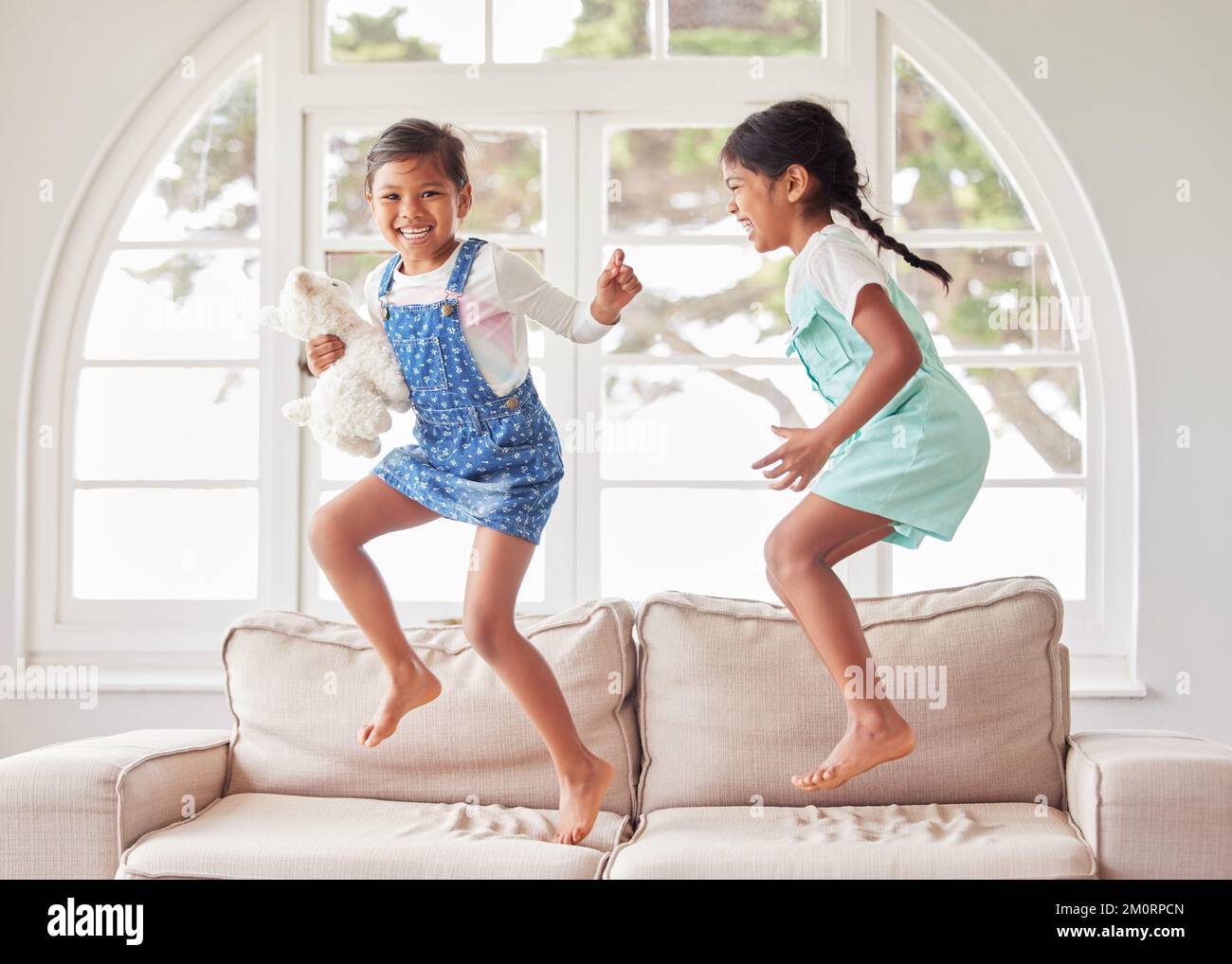 Due ragazze che saltano insieme sul divano. Due sorelle che si divertono e si guardano felici mentre ballano e suonano insieme a casa Foto Stock