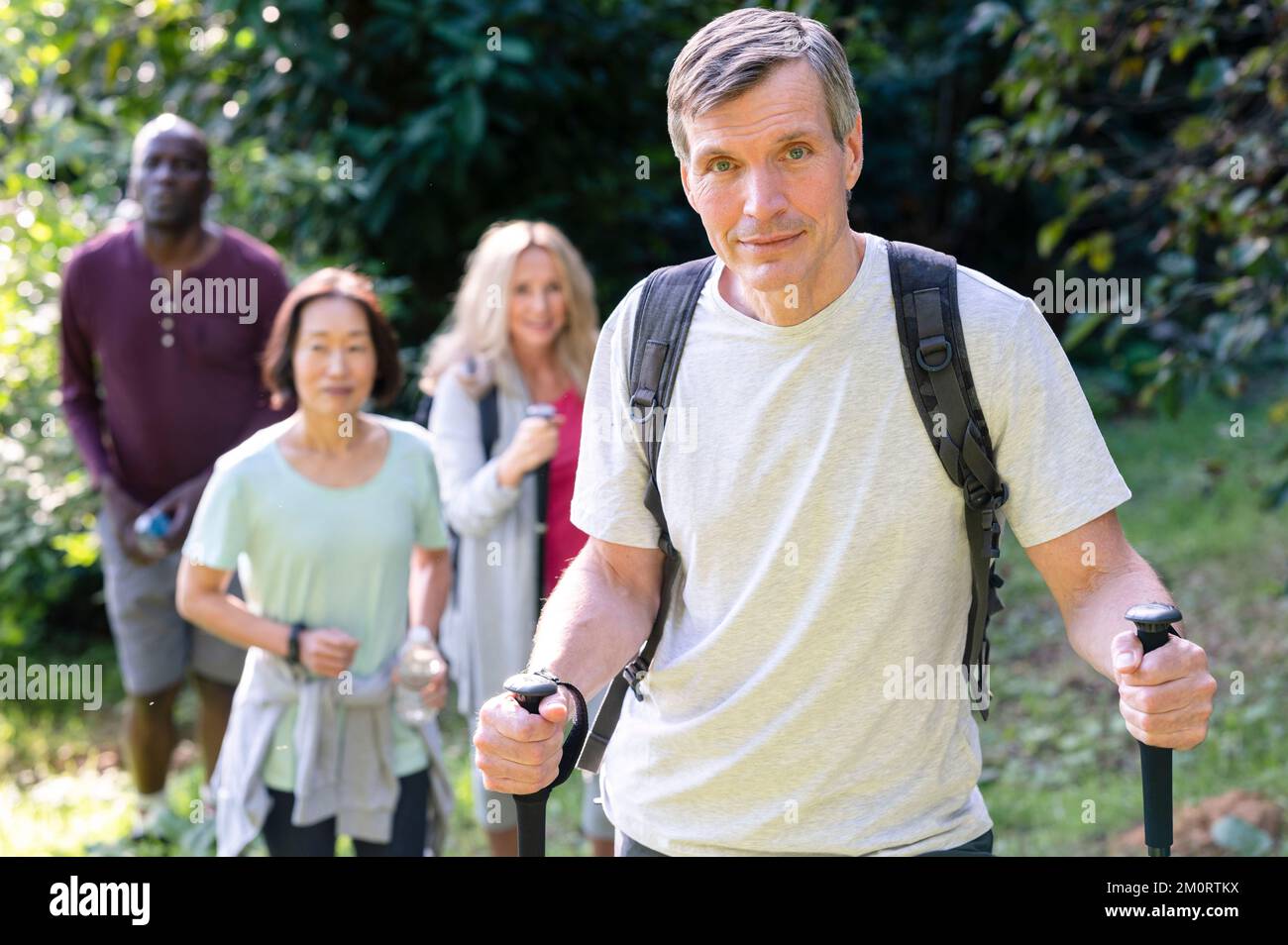 Uomo di mezza età dall'aspetto sano con bastoncini da trekking e zaini che camminano nelle strade secondarie locali con il suo variegato gruppo di amici Foto Stock