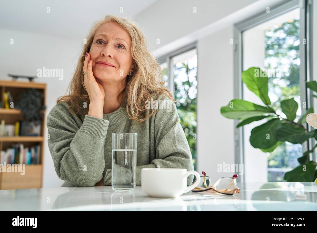 Donna anziana sorridente che guarda in alto mentre è seduta con la mano sul mento Foto Stock