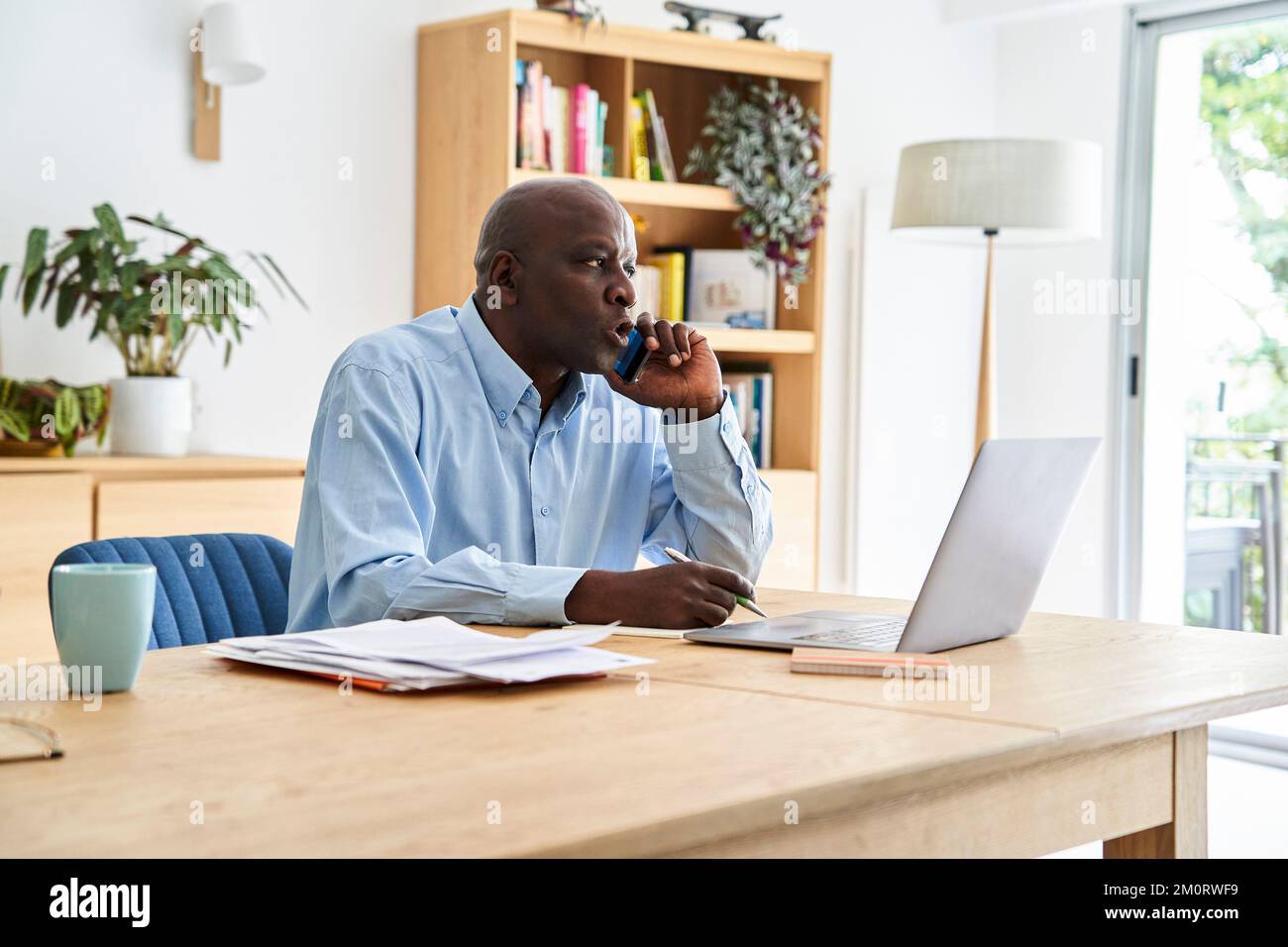 Uomo afro-americano di mezza età che effettua telefonate mentre lavora a casa con un computer portatile Foto Stock