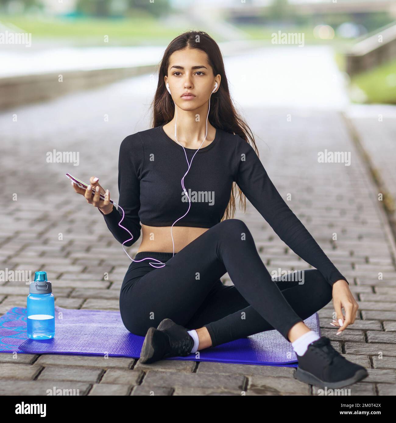 Bella giovane donna che conduce uno stile di vita sano, ascoltando la musica mentre si siede su un materassino ginnico, riposandosi dopo aver giocato gli sport sulla strada Foto Stock