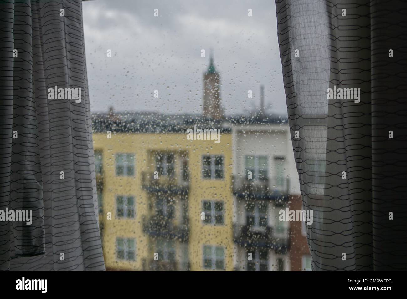 Le tende inquadrano una vista della città da una finestra coperta da gocce di pioggia in una giornata nuvolosa. Foto Stock