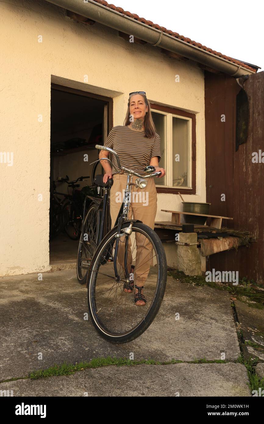 Migliore Ager femmina modello 50 Plus con invecchiamento bicicletta migliore. Frau im besten Alter ganz sportlich mit einem Fahrrad Oldtimer vor einem Schuppen. 50+, età Foto Stock