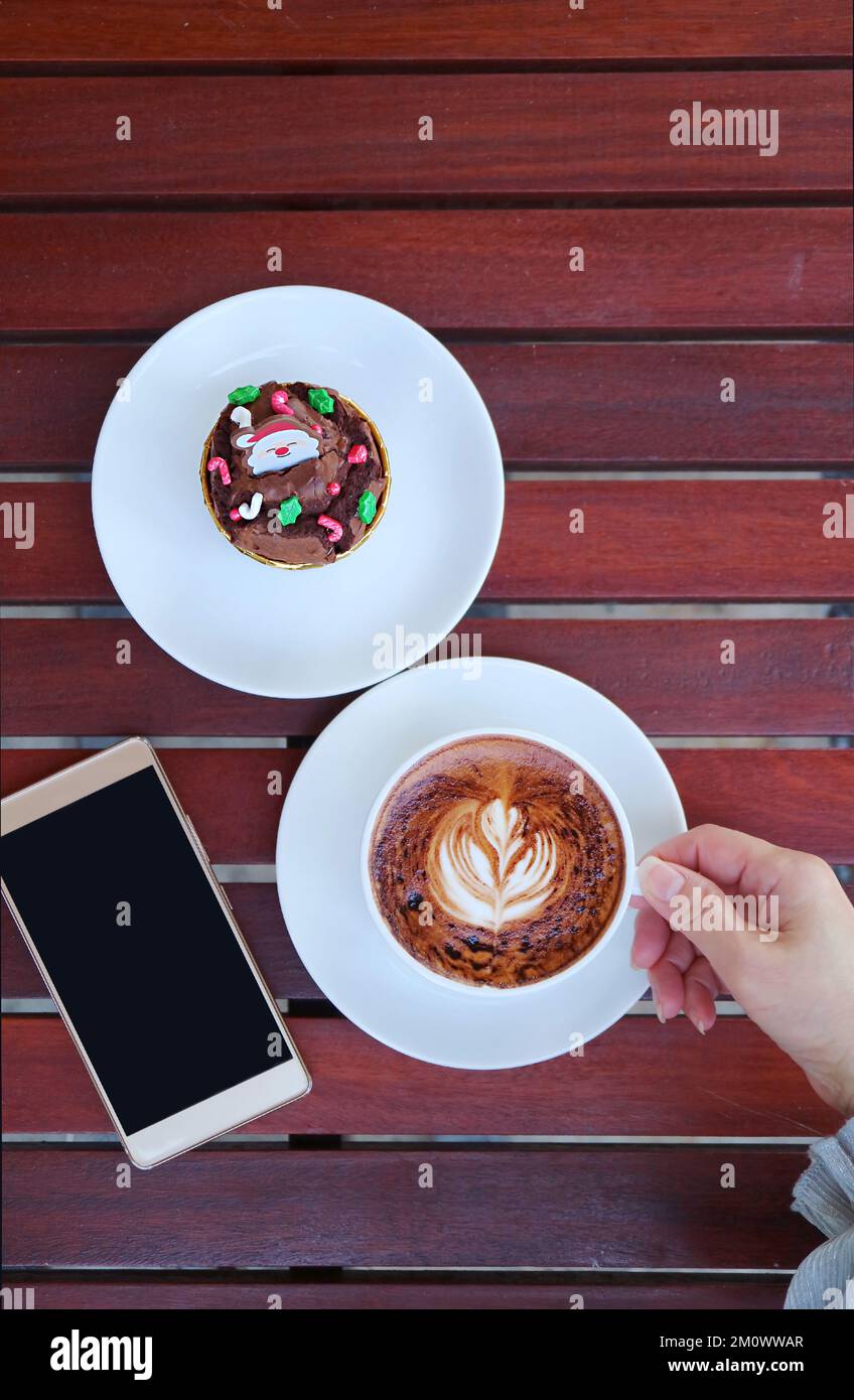 Tenere a mano una tazza di caffè caldo cappuccino accanto con un piatto di cioccolato di Natale Cupcake e smartphone Foto Stock