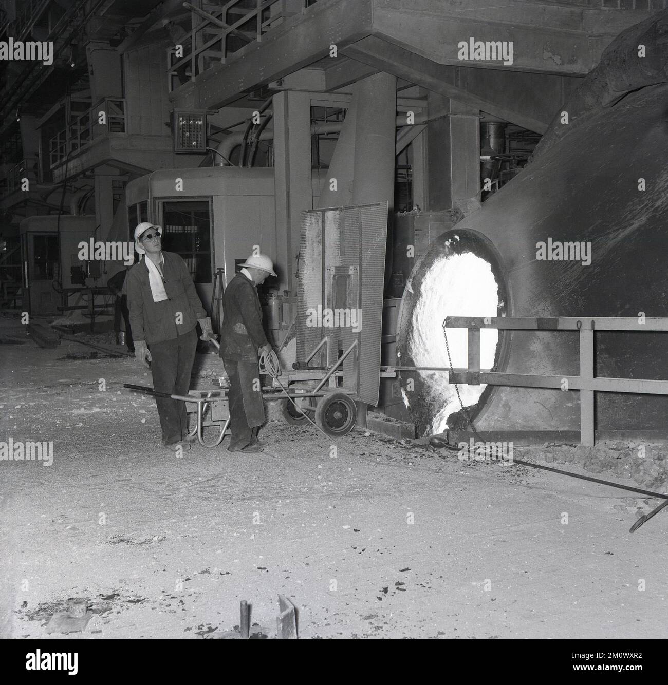 1950s, storico, acciaierie, due operai siderurgici in una fornace, in piedi dietro una guardia di protezione, uno che tiene una barra di acciaio nella fornace, Abbey Works, Port Talbot, Galles del Sud, Regno Unito. Foto Stock