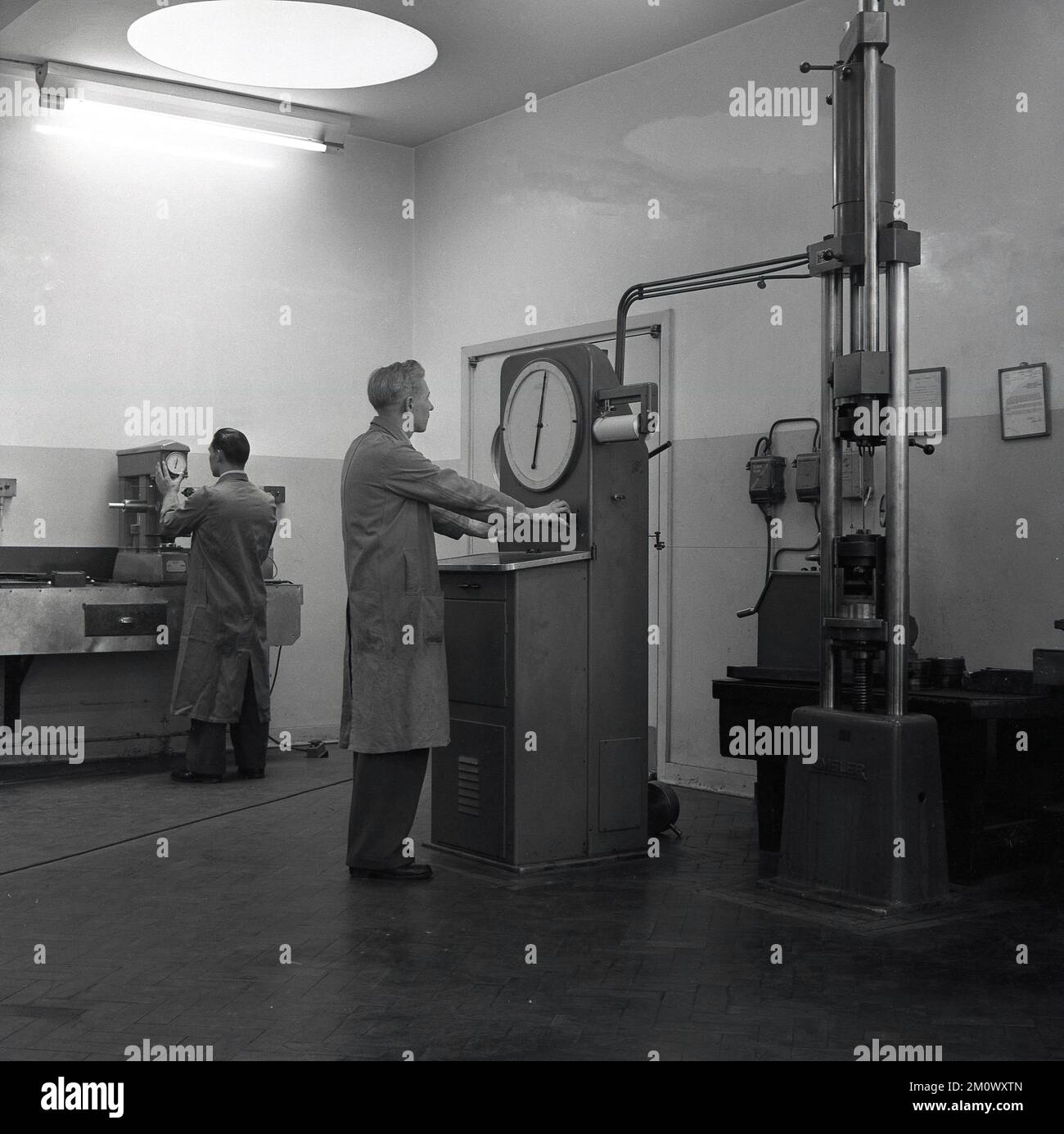 1950s, storico, acciaieria, una lunga operatrice rivestita all'interno che lavora con una macchina di controllo industriale con un grande quadrante presso Abbey Works, Port Talbot, Galles del Sud, UK. Una delle macchine viste è prodotta da Amsler, un'azienda svizzera che ha realizzato macchinari per la prova di lamiere. Foto Stock 1950s, storico, acciaieria, una lunga operatrice rivestita all'interno che lavora con una macchina di controllo industriale con un grande quadrante presso Abbey Works, Port Talbot, Galles del Sud, UK. Una delle macchine viste è prodotta da Amsler, un'azienda svizzera che ha realizzato macchinari per la prova di lamiere. Foto Stock