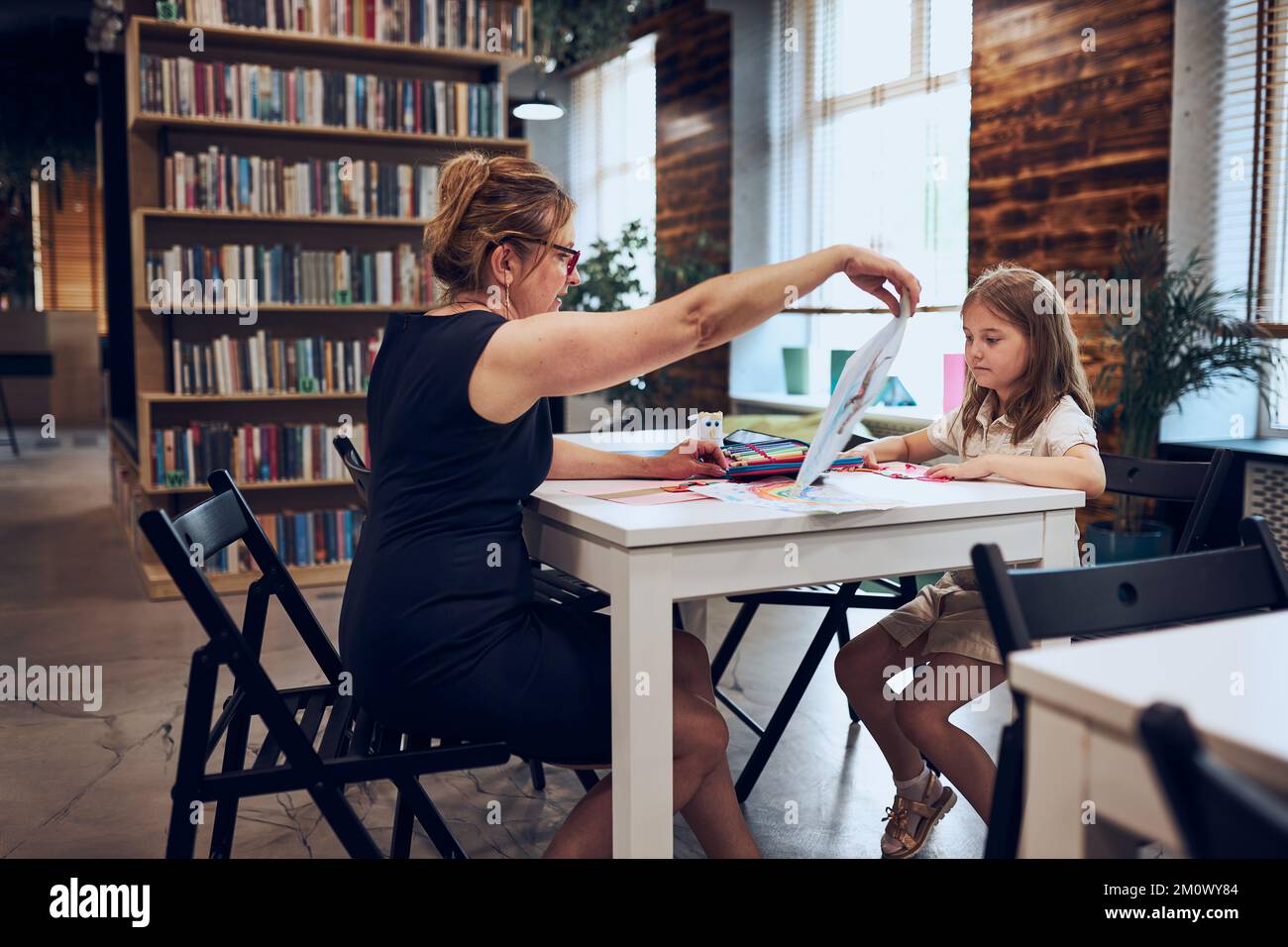 Insegnante che insegna scolastica durante le lezioni della scuola elementare. I bambini lavorano seduti alla scrivania nel club del postscuola. Ragazza disegno immagini. Imparando a. Foto Stock