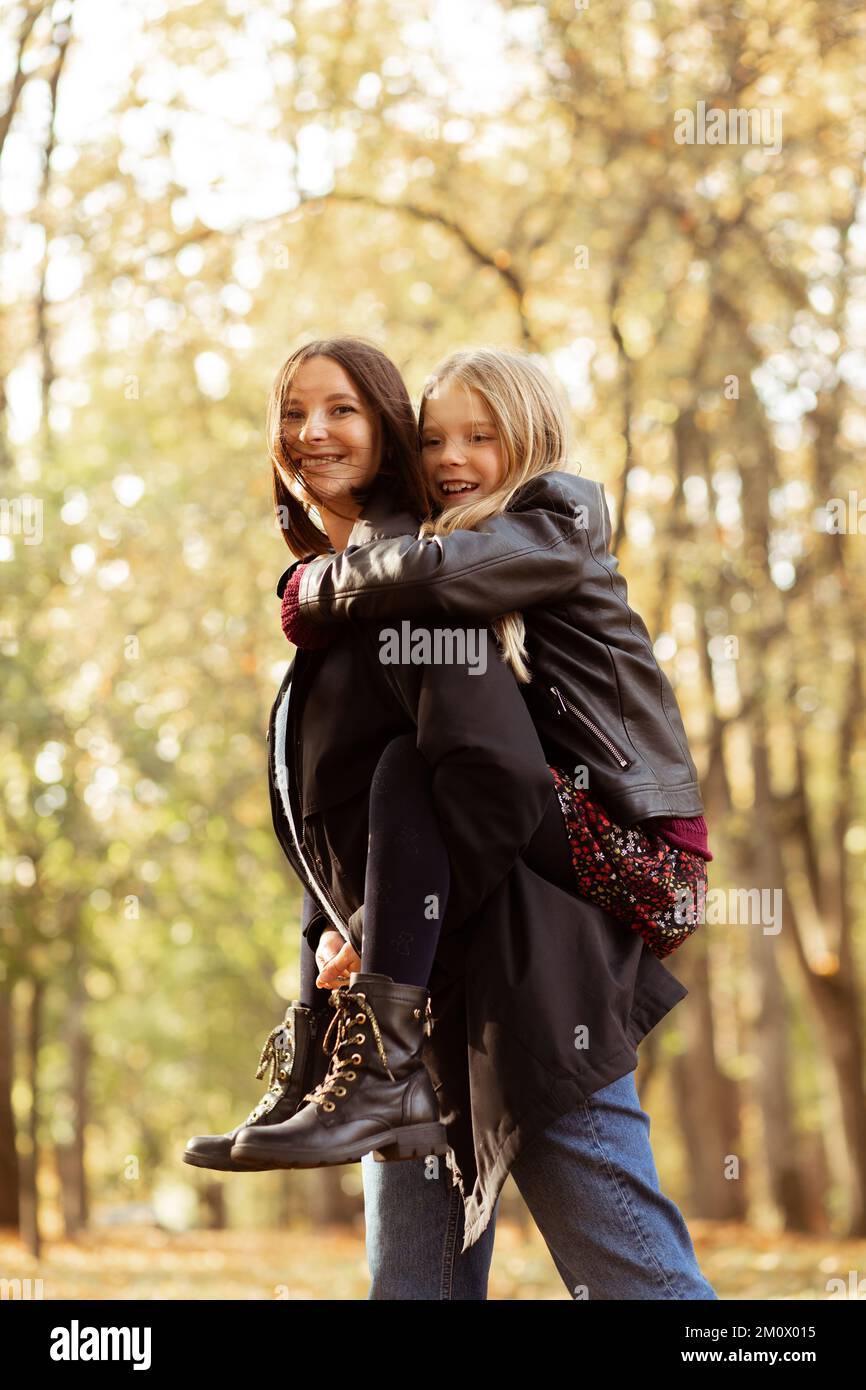 Allegro, allegro, positivo famiglia di madre che tiene la figlia sulla schiena, camminando nella foresta d'autunno dorato. Adozione da parte dei bambini Foto Stock