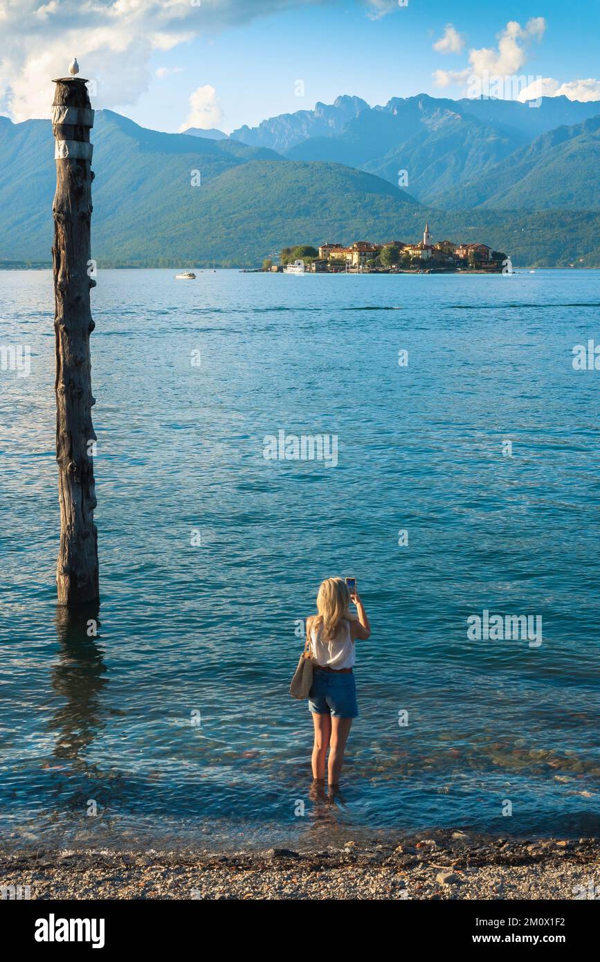 Vacanza femminile, vista posteriore in estate di una giovane donna in piedi sul Lago maggiore e scattando una foto di Isola dei pescatori, Piemonte, Italia Foto Stock