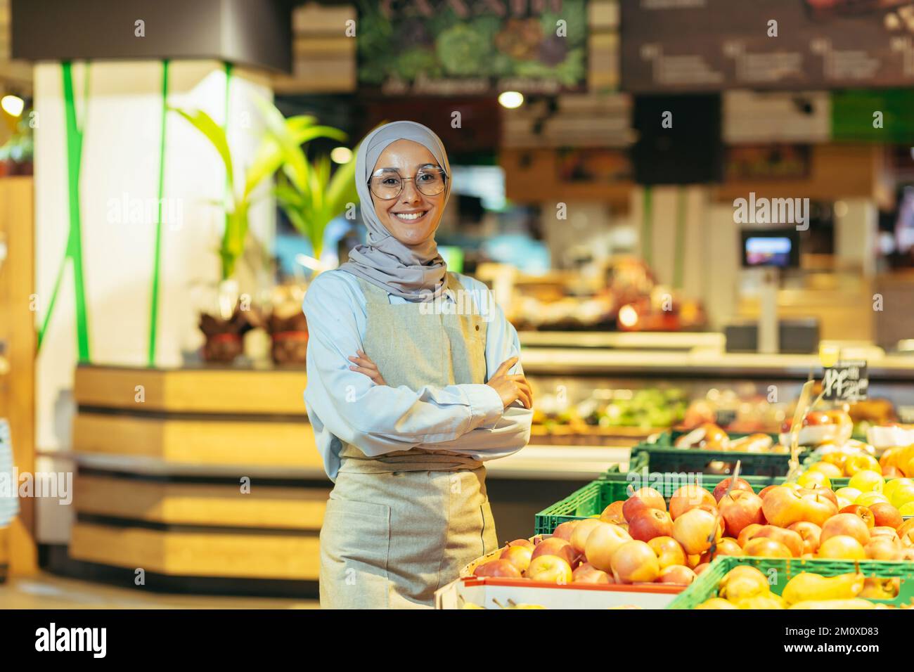 Ritratto di una bella giovane donna musulmana in un hijab, che lavora in un supermercato. Si trova nel reparto di verdure e frutta, le attraversa le braccia, guarda la macchina fotografica. Sorride. Foto Stock