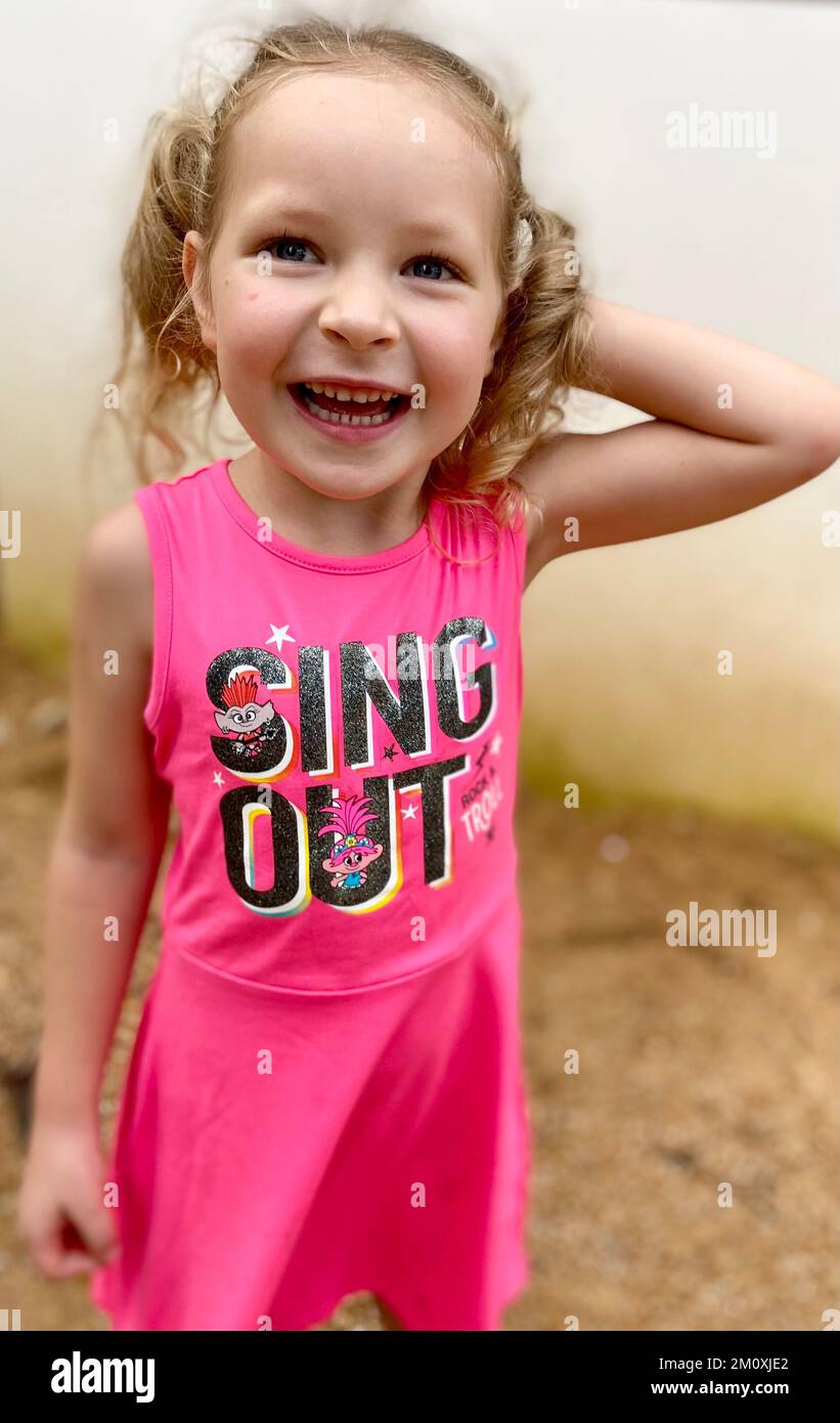 Una ragazza caucasica bionda di 5 anni con pigtail in un abito rosa con un film Trolls stampa sorridente Foto Stock