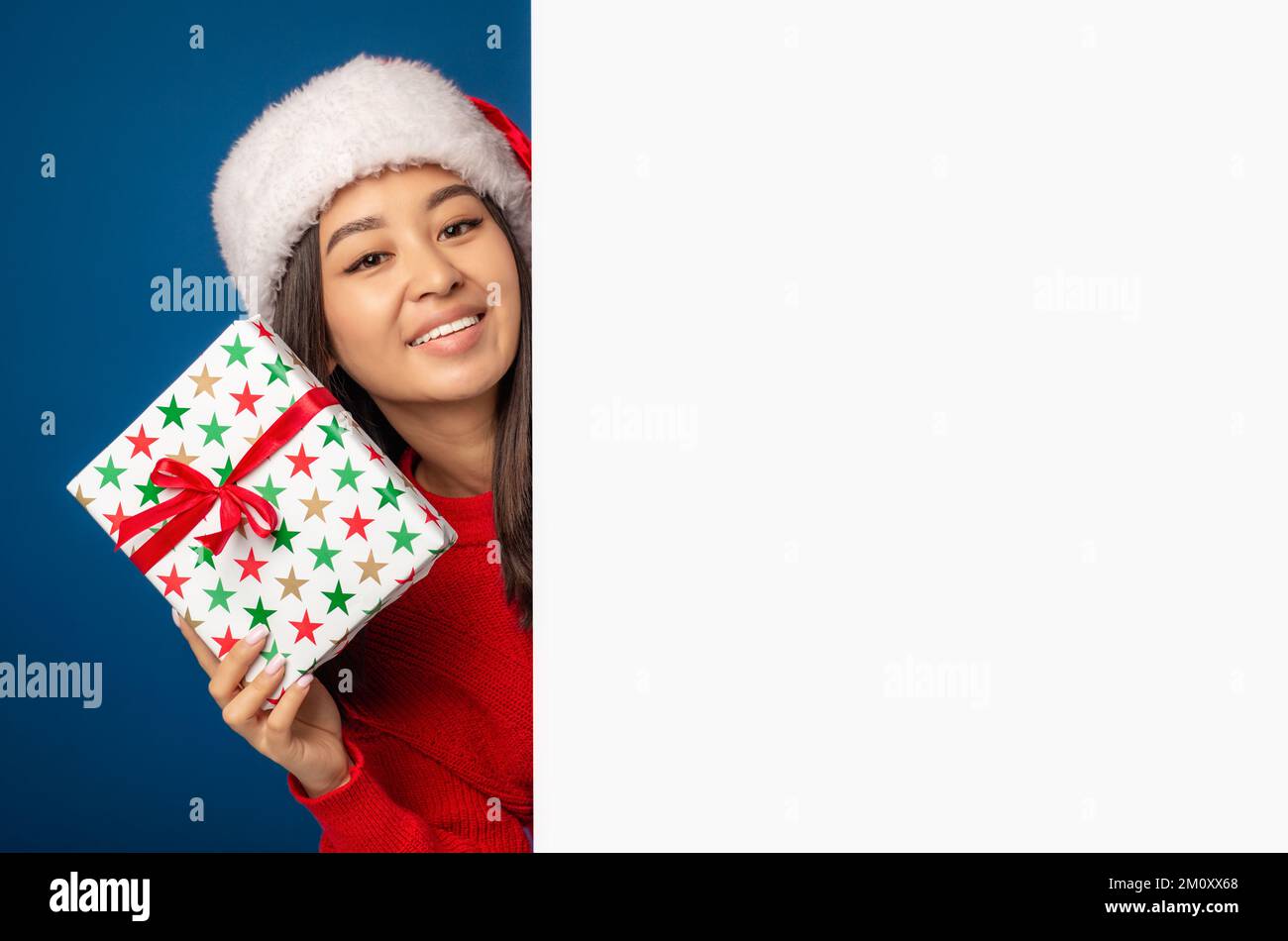 Donna sorpresa positiva in cappello di babbo natale con il regalo di Natale che si scopano fuori dal bordo bianco vuoto, concetto del regalo di Natale Foto Stock
