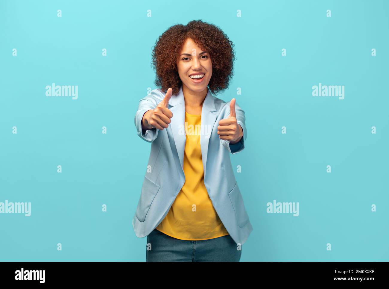 Donna afro-americana felice con capelli ricci in giacca approvando fare gesto positivo con la mano su sfondo blu Foto Stock