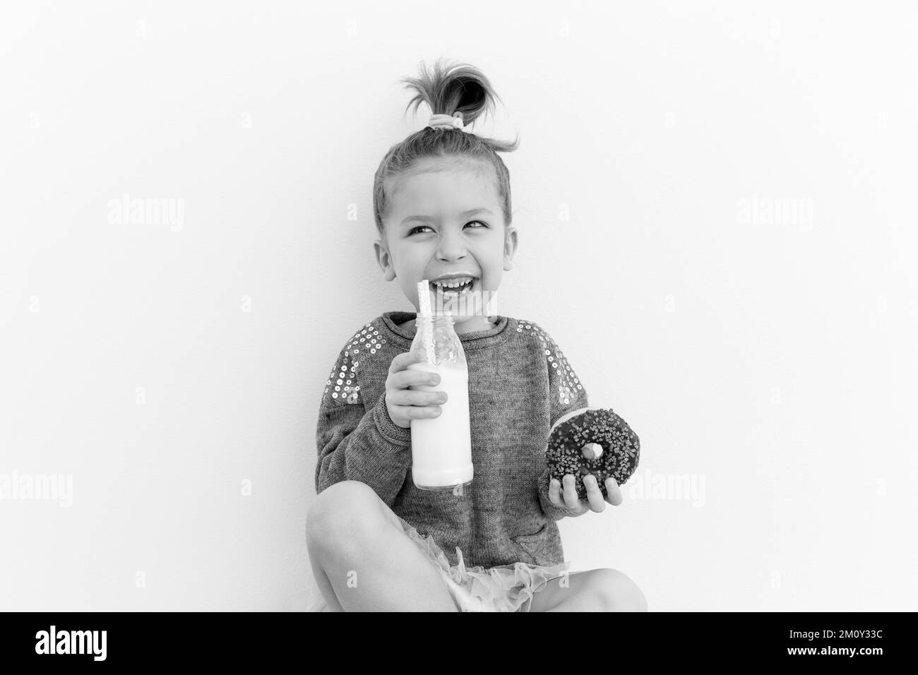 La bambina adorabile sta mangiando ciambella al cioccolato con una bottiglia di latte. Divertendosi, cibo allegro. Il concetto di amante dei dolci. Foto in bianco e nero Foto Stock
