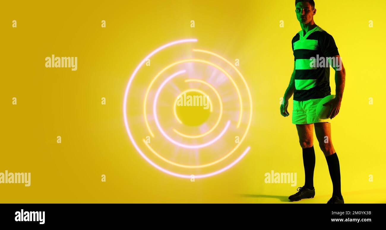 Tutta la lunghezza del giocatore di rugby caucasico con palla in piedi da un motivo circolare illuminato, copia spazio. Giallo, composito, sport, competizione, gioco, Foto Stock