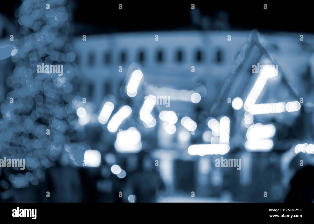 Sfondo sfocato. La gente cammina nella piazza della città la sera d'inverno. Sagome di persone che camminano vicino a case decorate illuminazione luminosa. Luce bianca spot bokeh da luci di casa incandescente colore blu Foto Stock