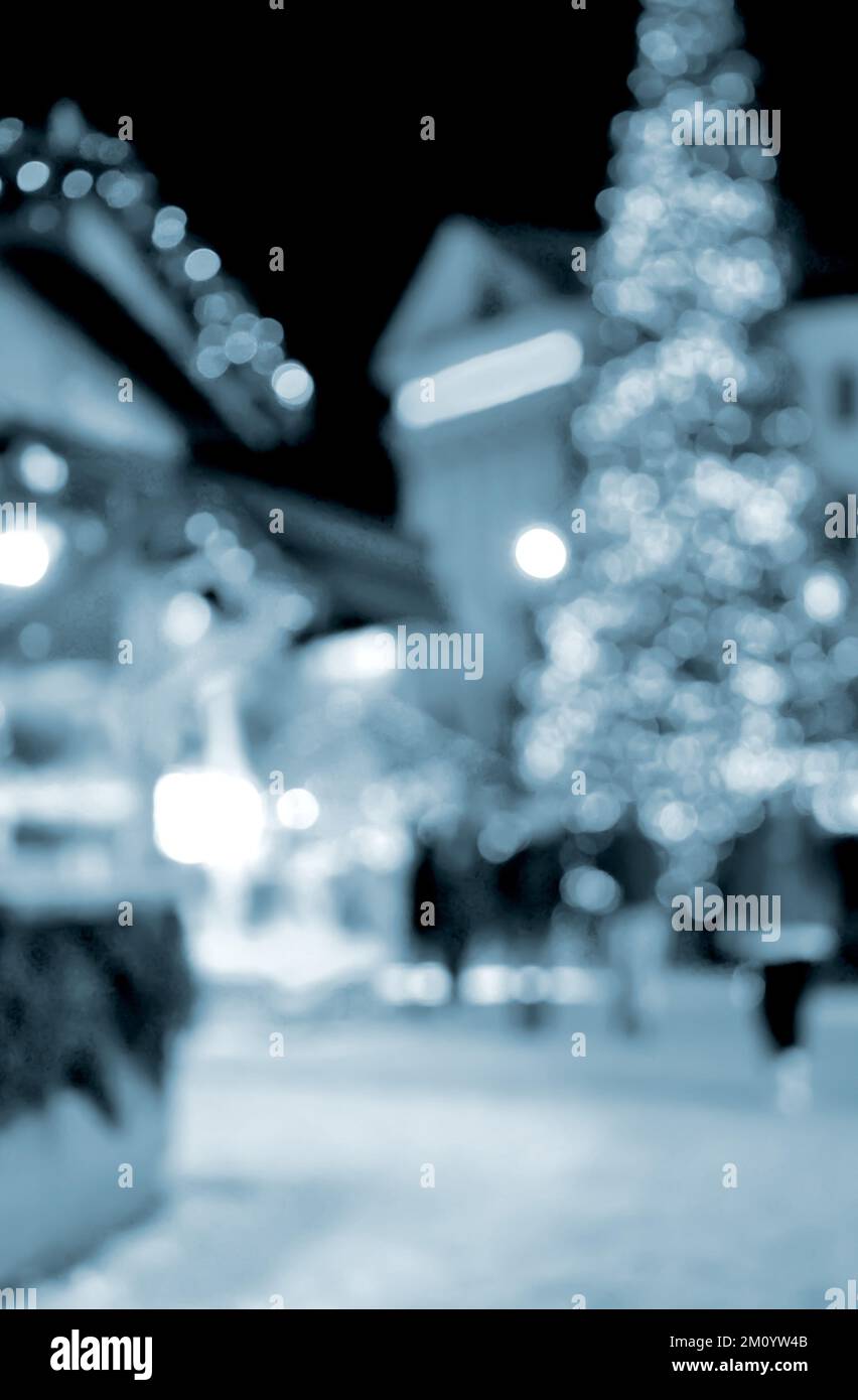 Sfondo sfocato. Albero di Natale di Capodanno decorato giallo ghirlande illuminazione, piccole case di legno ghirlande sul tetto e sagome di persone che camminano lungo la strada della città. Colore blu Foto Stock