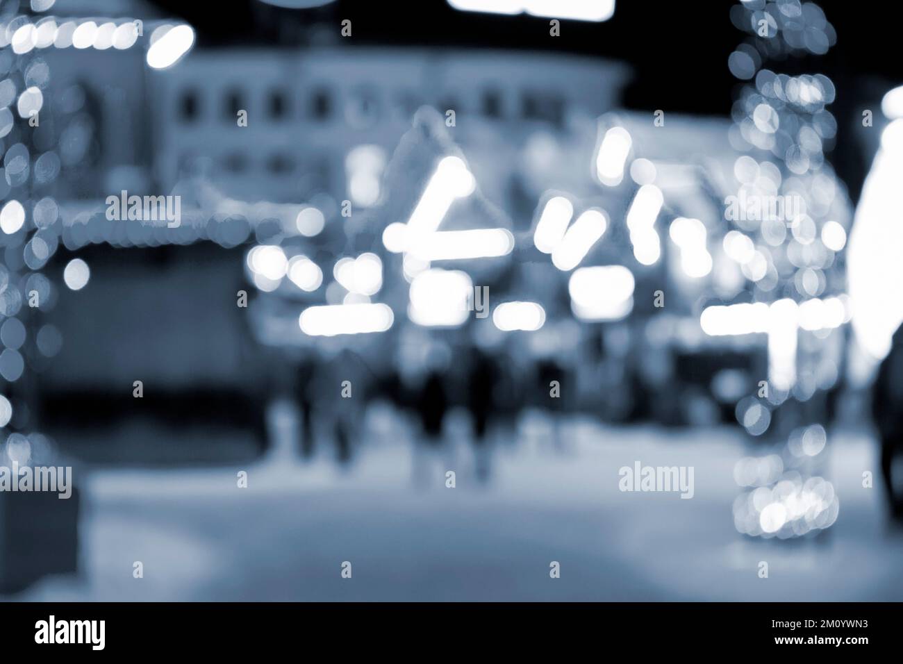 Sfondo sfocato. La gente cammina in città la notte d'inverno. Silhouette nere di persone che camminano vicino a case decorate illuminazione luminosa. Luce bianca spot bokeh da luci di casa incandescente colore blu Foto Stock