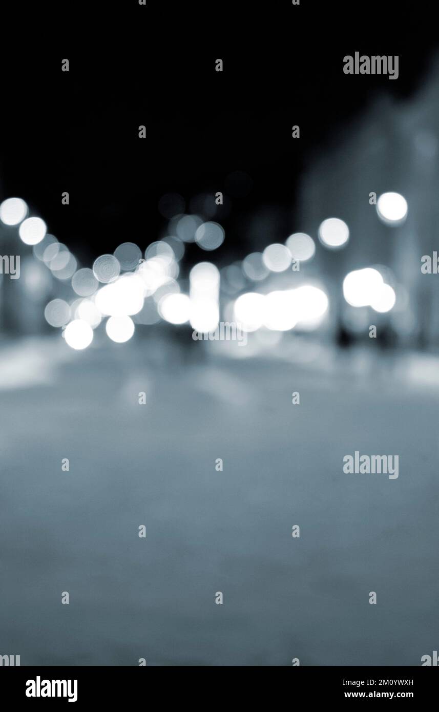 Sfondo sfocato. Notte città. Silhouette sfocata degli edifici, macchie bokeh di lanterne luminose, silhouette sfocate di persone che camminano per strada di sera. Piazza della città, cielo notturno nero di colore blu Foto Stock