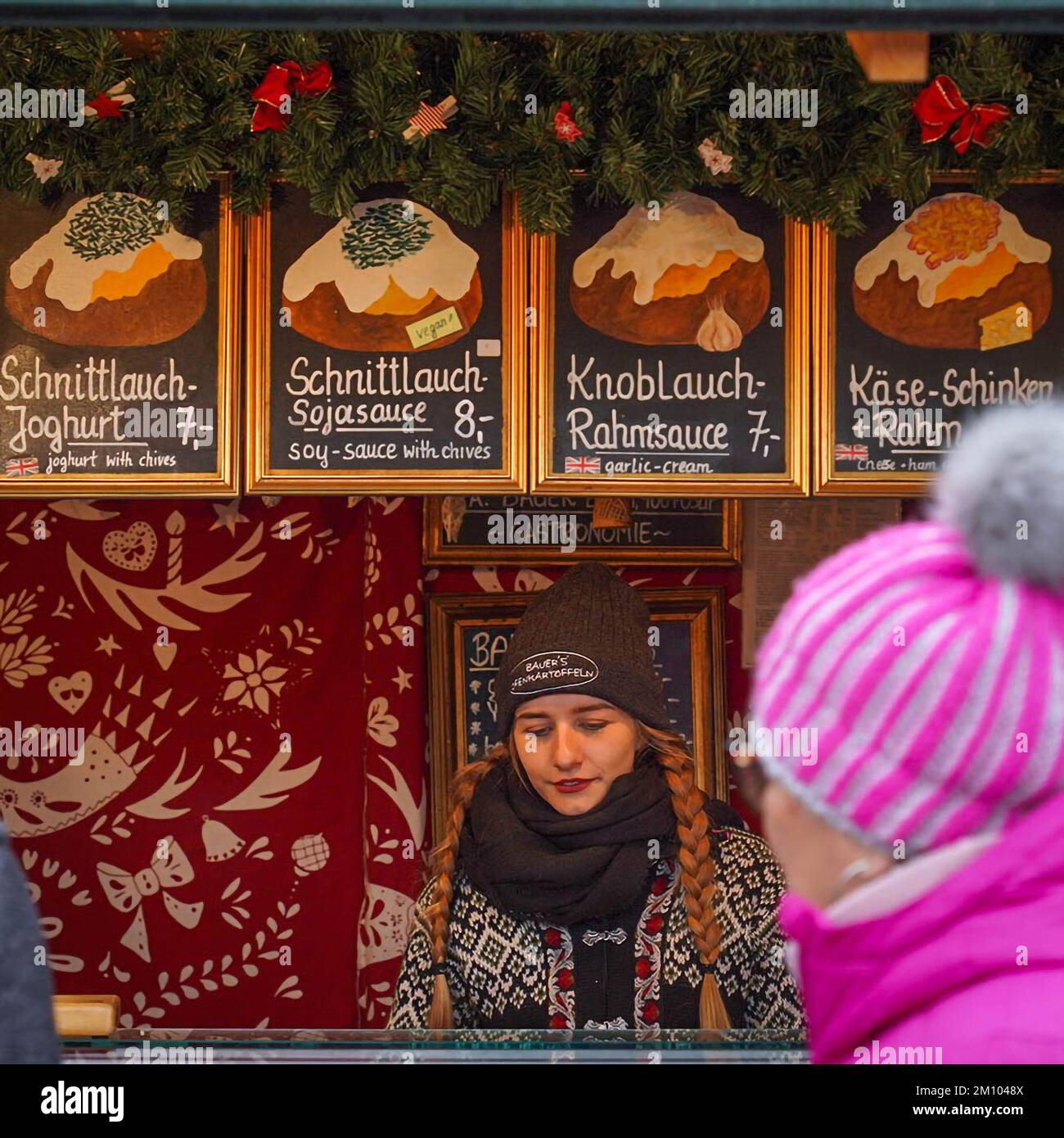 Una giovane donna venditore si trova dietro il bancone con un negozio nel chiosco del mercato di Natale, che vende patate al forno Foto Stock
