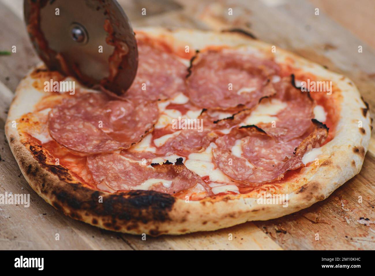 Tagliare la pizza calda con salsa di pomodoro, mozzarella e salame con un taglierino su un asse di legno, affumicato, primo piano Foto Stock