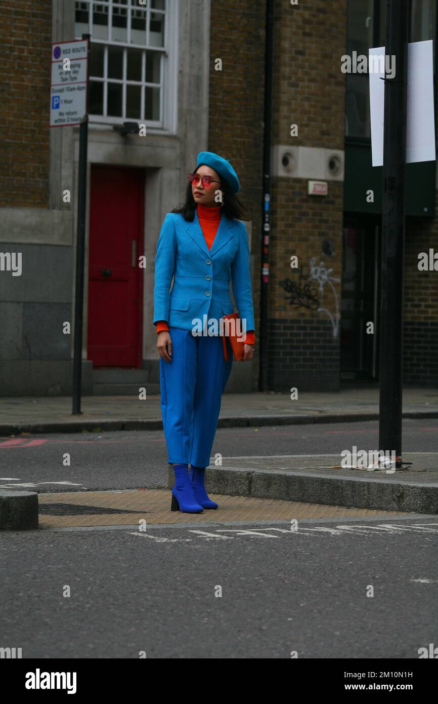 Elegante donna in berretto blu, stivali blu, pantaloni blu, giacca blu, dolcevita arancione, Occhiali da sole e una pochette arancione su una strada londinese Foto Stock