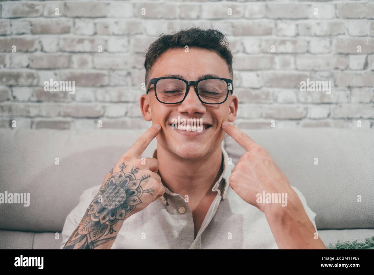 Un giovane tatuato sorrise e guardò la telecamera mentre si divertiva a toccare e a indicare il suo sorriso. Ritratto dell'adolescente che indossa gli occhiali. Foto Stock