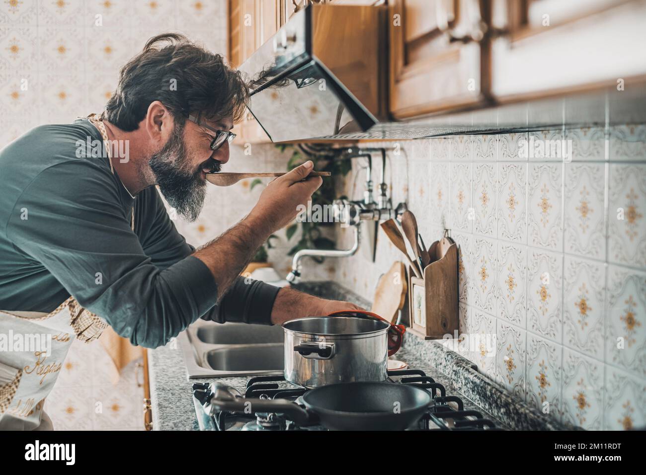 L'uomo a casa assaggia il cibo dalla pentola con un cucchiaio da tavola. Persone in cucina per preparare la cena. Un maschio maturo che cucina da solo. Persona di stile di vita reale che prova il gusto e la preparazione del cuoco. Lavoro del marito Foto Stock