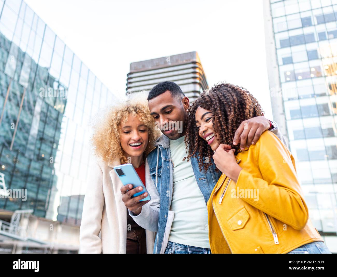 Da sotto allegro giovane multirazziale Signore in abiti eleganti sorridendo felicemente e abbracciando positivo maschio migliore amico utilizzando il telefono cellulare sulla piazza della città Foto Stock