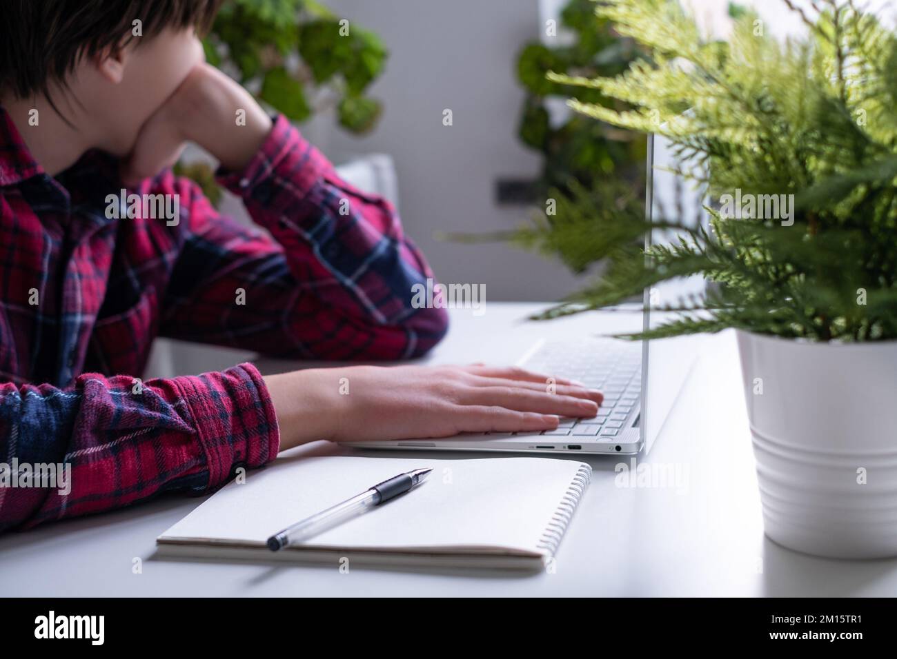 Il ragazzo sta lavorando su un computer portatile. Scolaro stanco si appoggia sulle mani, burnout, stress con mal di testa a tavola. Formazione online, home Office. Poco profondo Foto Stock