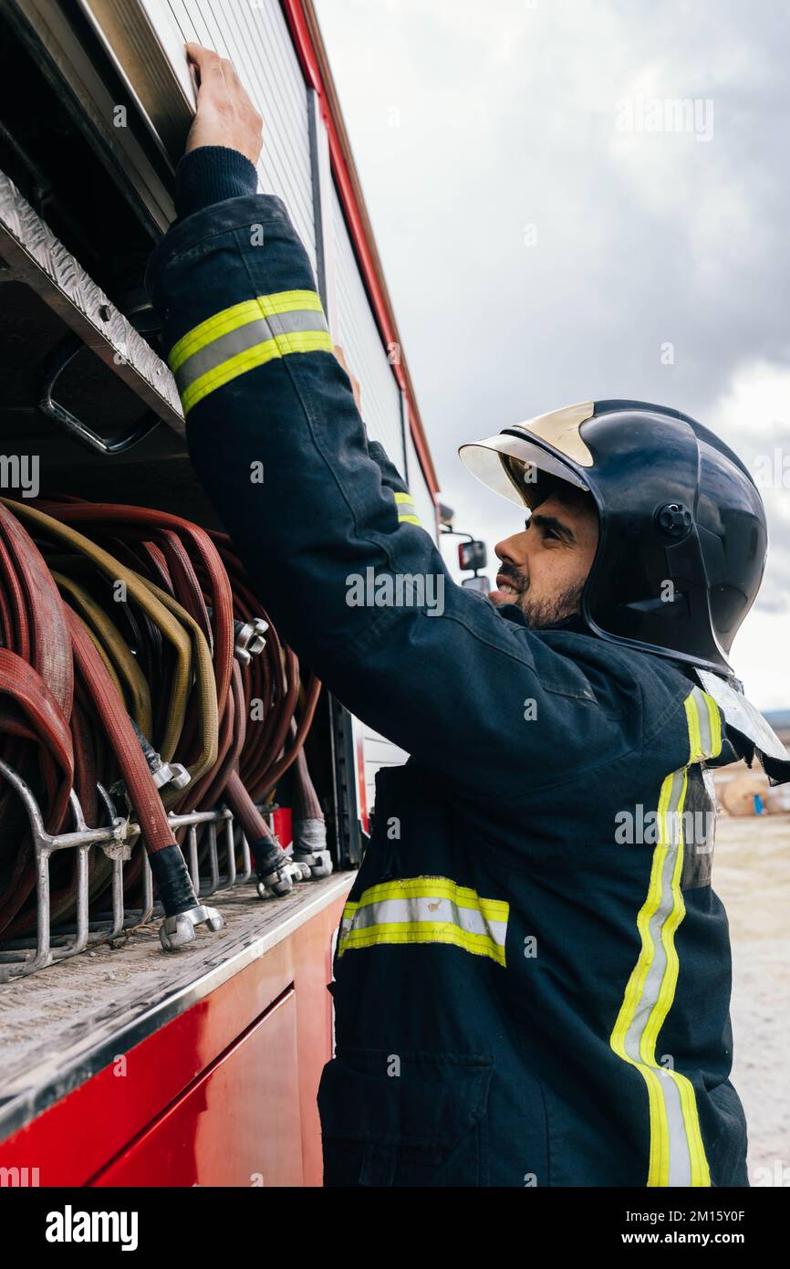 Positivo focalizzato ispanico pompiere sorridente e chiudente tubo vano di fuoco contro cielo nuvoloso sulla stazione Foto Stock