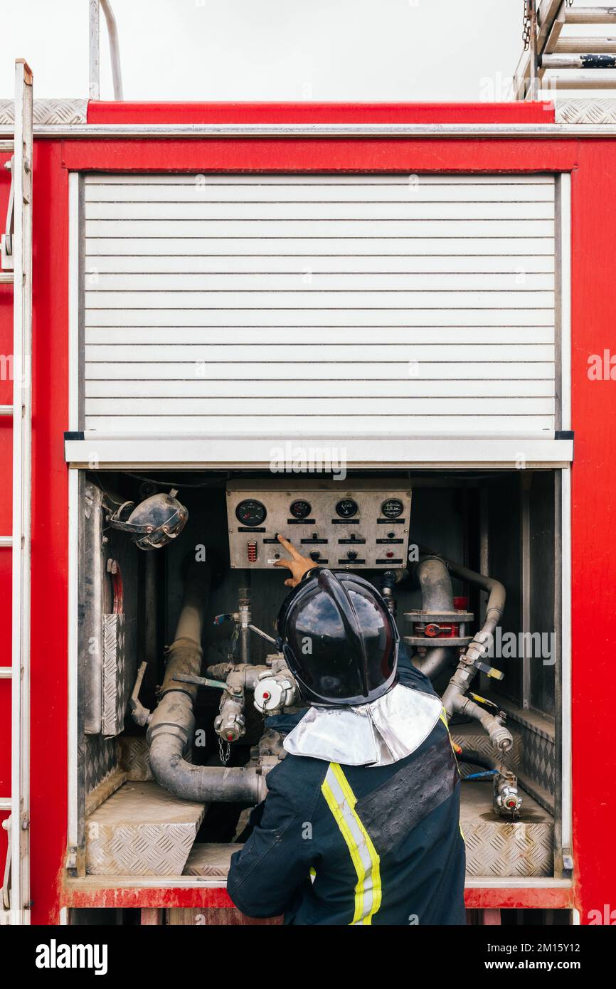 Vista posteriore di un pompiere irriconoscibile che indica il pannello di controllo ed esamina i display sulla pompa dell'acqua del motopompa antincendio durante il lavoro Foto Stock