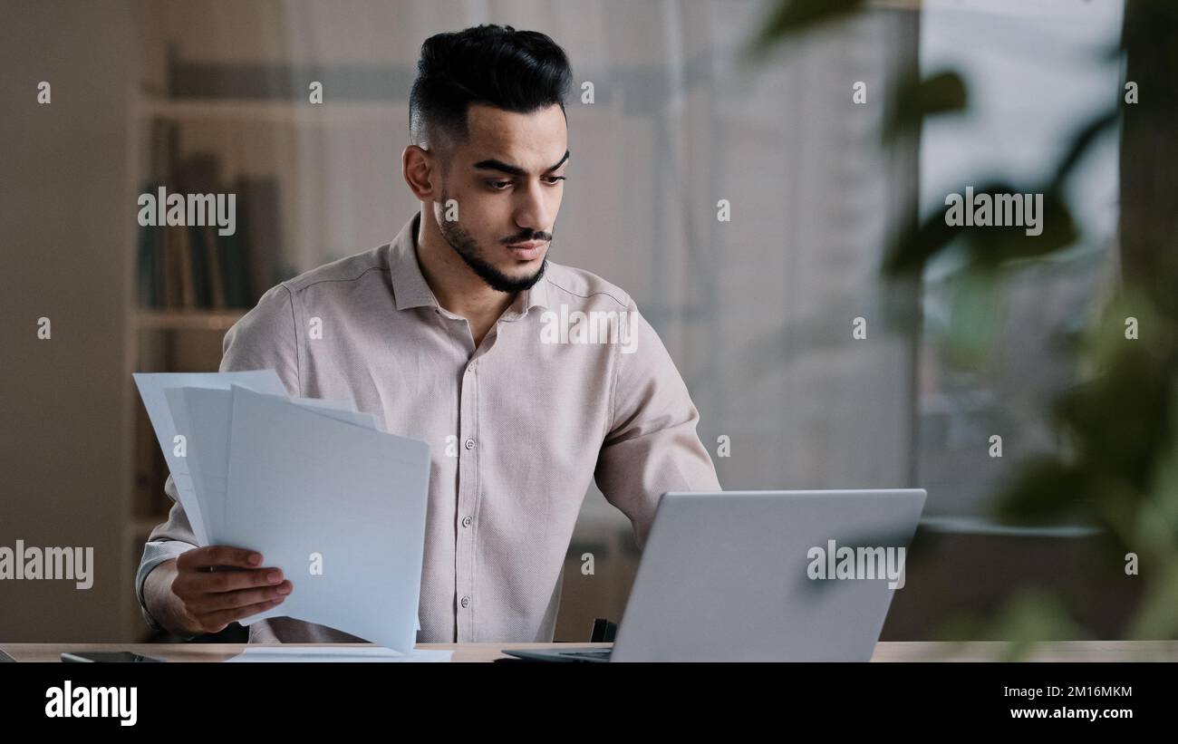 Serio giovane uomo d'affari arabo maschio professionista lavoratore dipendente tipo su tastiera computer copia testo cartaceo in forma elettronica focalizzato uomo ispanico Foto Stock