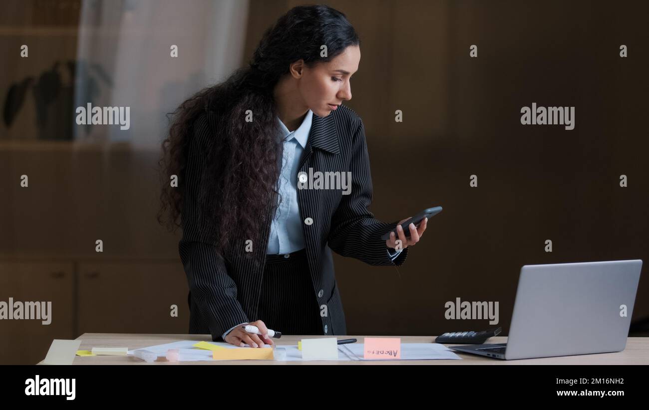 Donna di affari del progettista arabo del dipendente concentrata che si trova alla scrivania dell'ufficio usando il telefono mobile che controlla le informazioni che scrivono le attività sulle note adesive Foto Stock