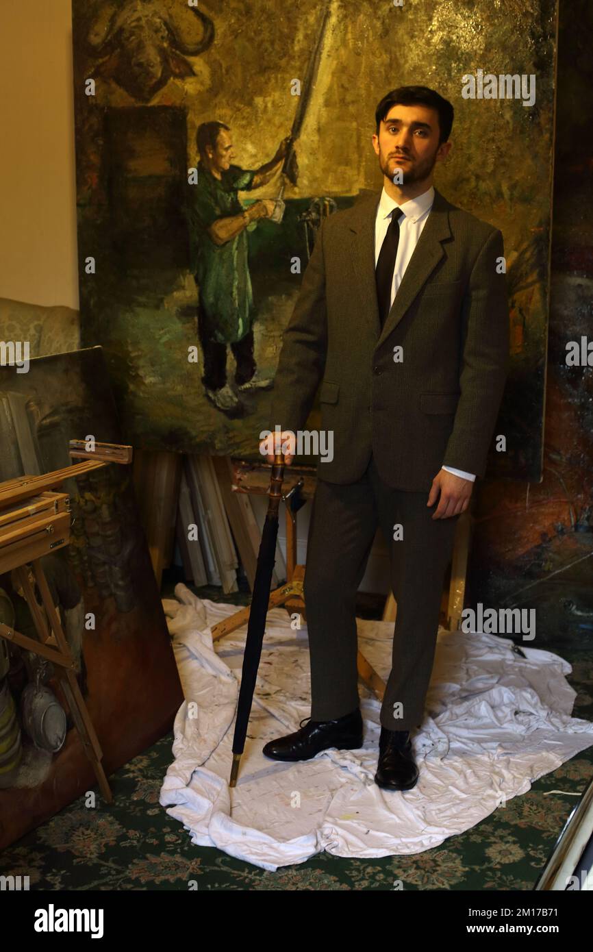 Uomo elegante vestito in tuta e punta che tiene un ombrello in uno studio d'artista con pittura ad olio sullo sfondo. Foto Stock