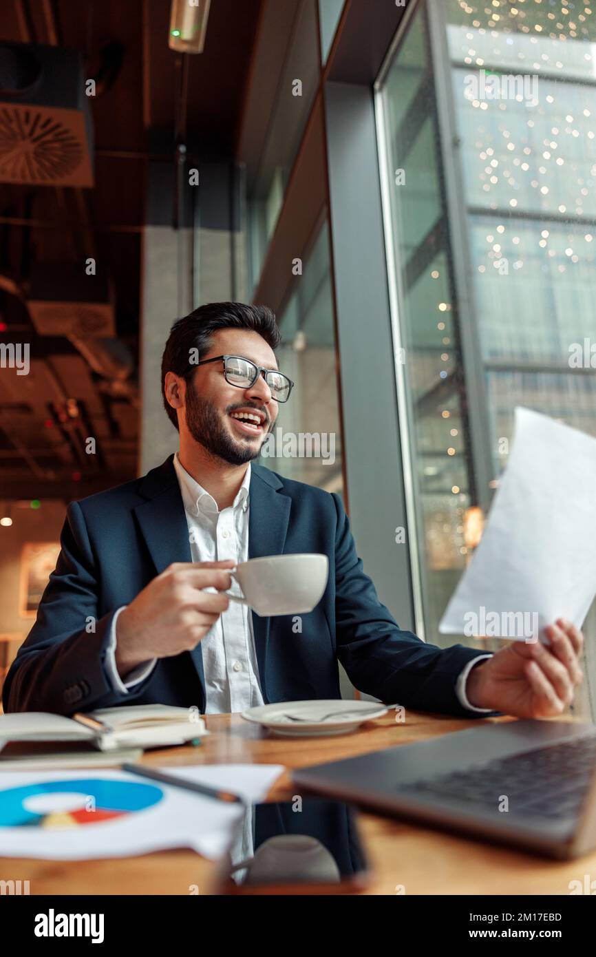 Uomo d'affari indiano sorridente che beve caffè e lavora con i documenti nel caffè Foto Stock